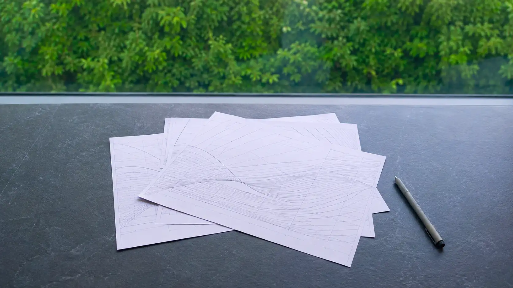 Mehrere Papierbögen mit gezeichneten Kurven fächrig auf einer dunklen Steinplatte, ein Stift daneben