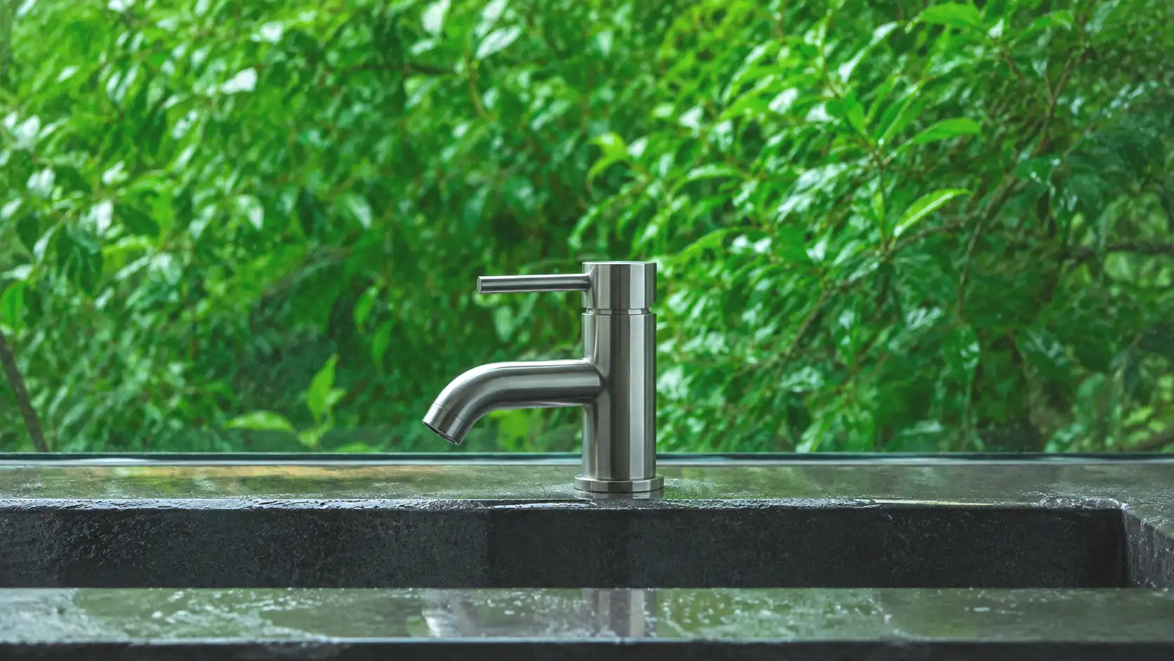 Badarmatur aus gebürstetem Edelstahl an einem Becken aus nassem dunklem Stein, dahinter dichtes Laub