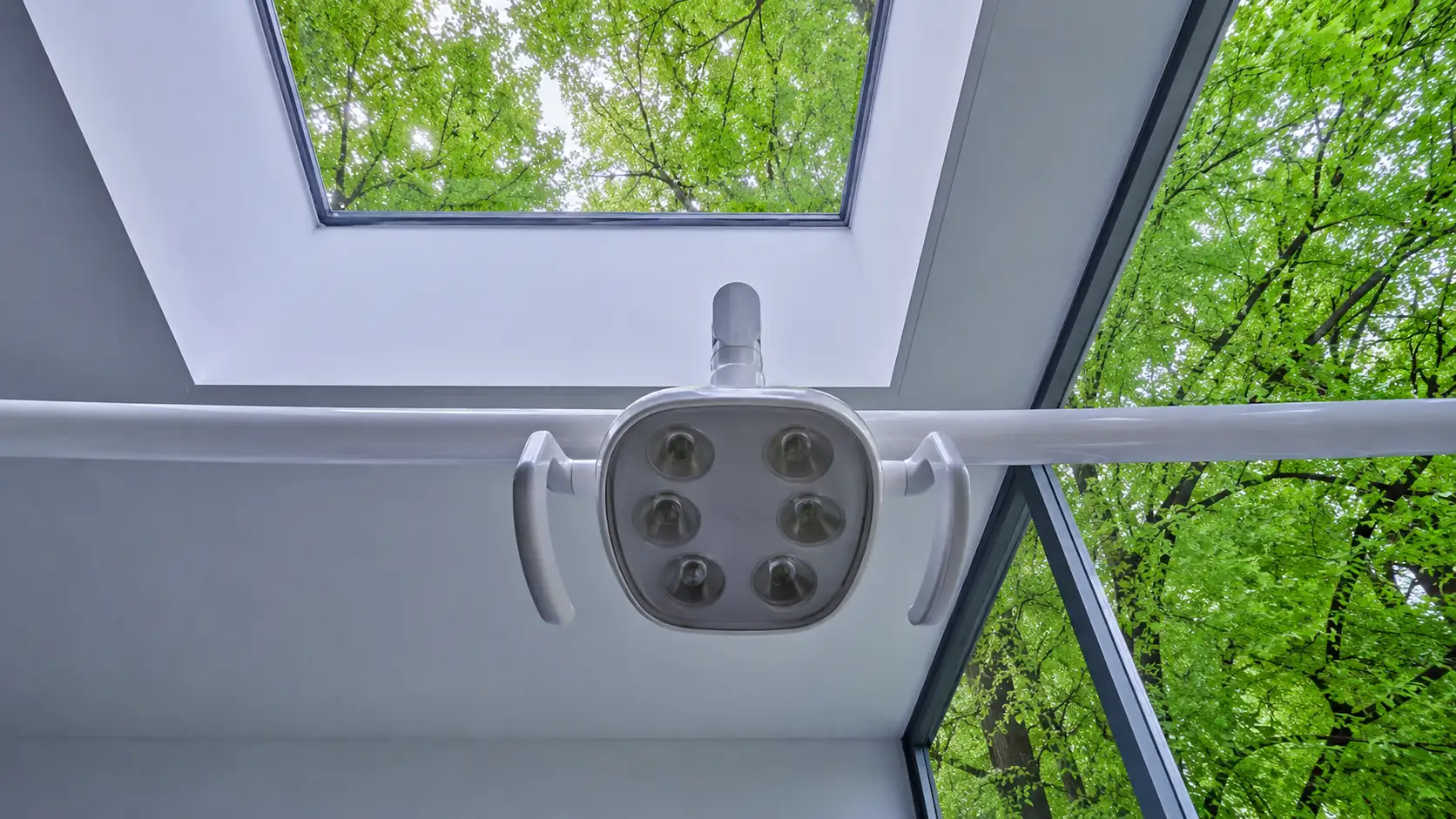 Ausgeschaltete zahnärztliche Behandlungsleuchte von unten gesehen, dahinter ein Dachfenster mit Baumkronen